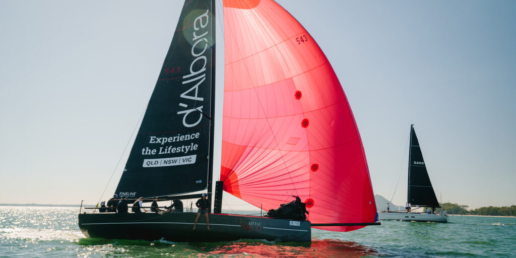 Sailing race on open water with a bright red spinnaker catching wind beside a dark sail with white text.