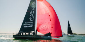 Sailing race on open water with a bright red spinnaker catching wind beside a dark sail with white text.